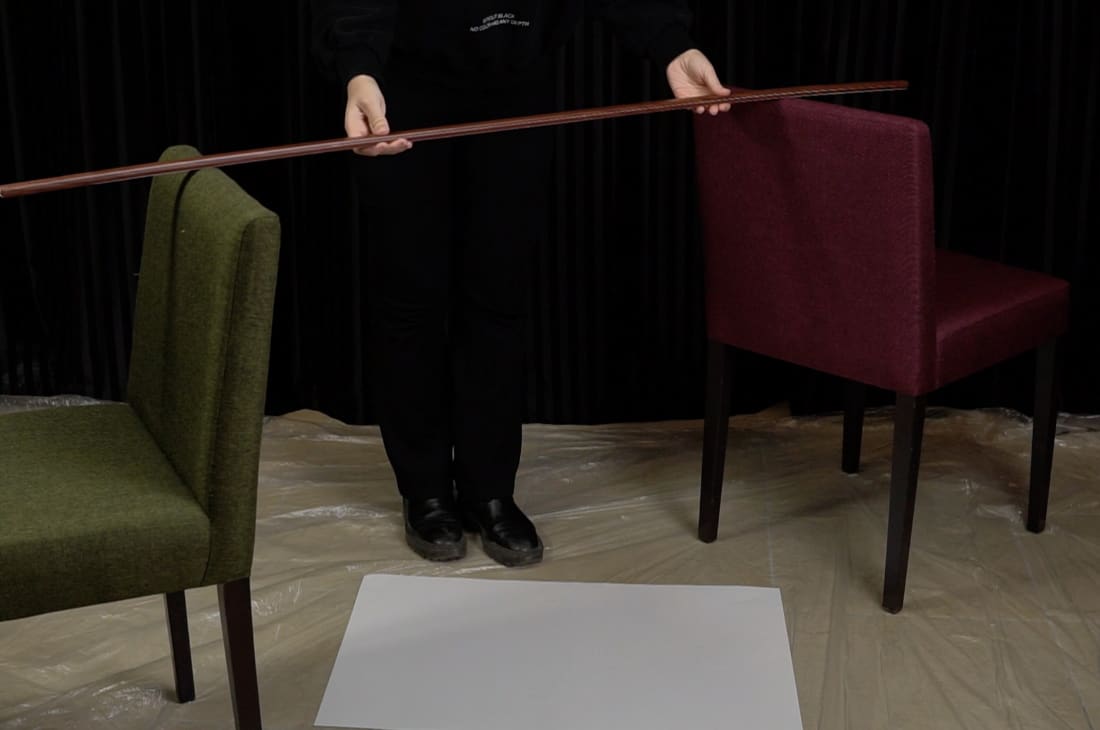 İki sandalyeyi resim kağıdının etrafına karşılıklı olarak yerleştir ve sopayı da sandalyelerin üzerine yerleştir.